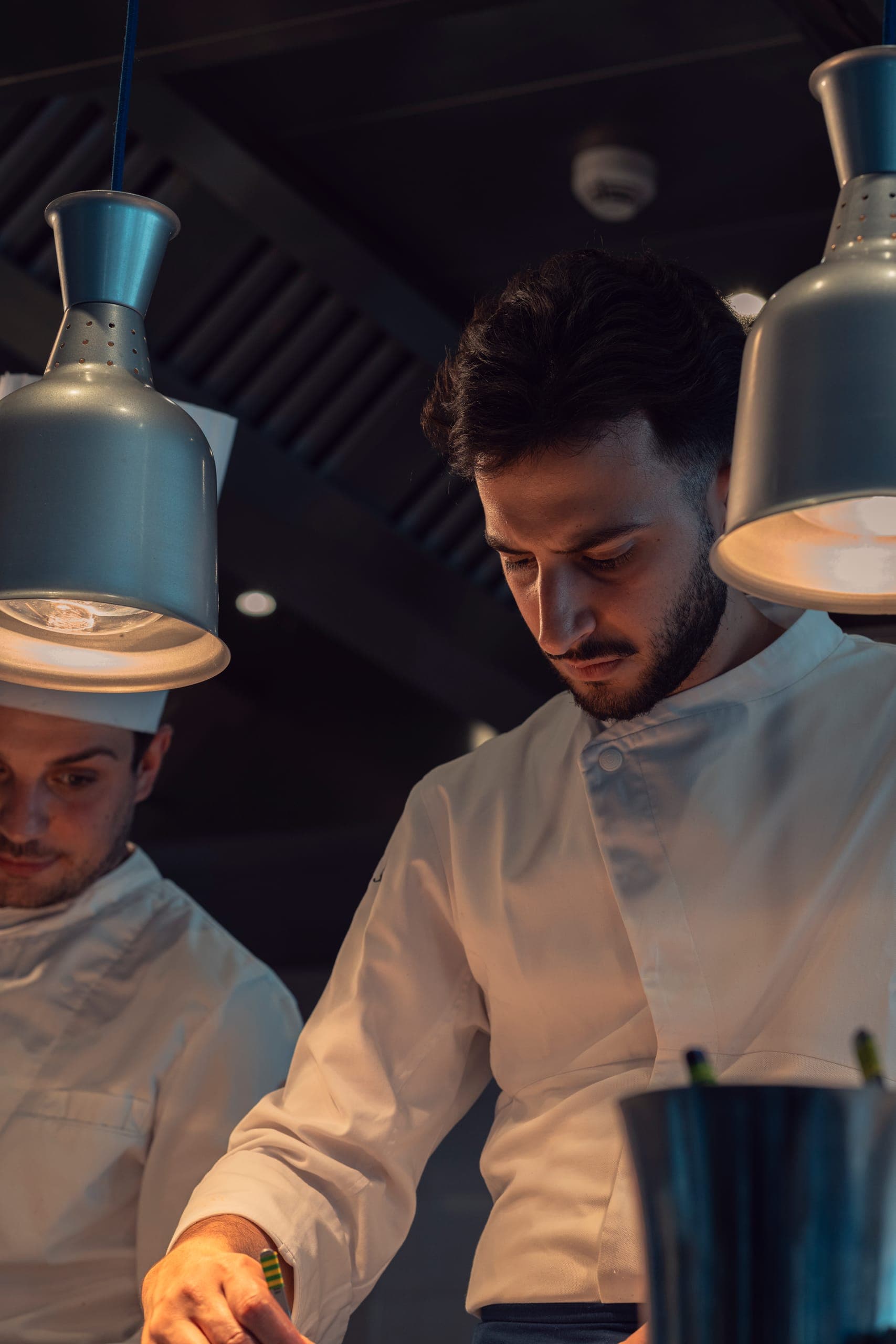 Due cuochi al passe, sotto le lampade calde del servizio.